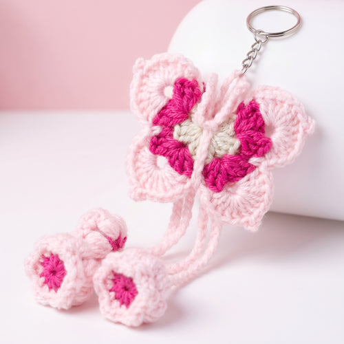 A pink and magenta crocheted butterfly keychain with two matching flower charms, displayed on a white cylindrical stand against a soft pink and white background.