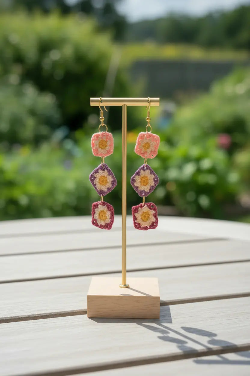 A pair of handmade dangle earrings displayed on a small gold stand on a wooden table, with a blurred garden background.