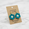 A pair of handmade crocheted flower earrings in teal and white, hanging from gold hooks on a brown kraft display card that reads 'handmade with heart', set against a light marble background.