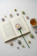 A purple and yellow knit flower bookmark with a long green stem rests on an open book on a white fabric surface, surrounded by small dried flowers and a cup of tea.