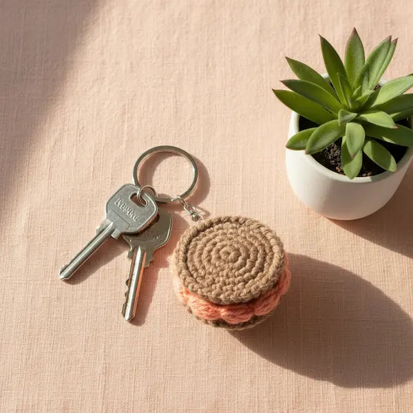 A crocheted keychain shaped like a sandwich cookie in beige and peach, attached to a metal key ring with two keys, placed on a pink textured surface next to a small potted succulent.
