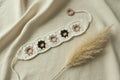 A crocheted beaded bracelet laid flat on a cream fabric background with a dried pampas grass stem. The bracelet is off-white with five circular beads: three dusty pink and two dark brown.