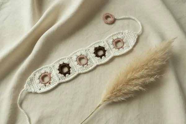 A crocheted beaded bracelet laid flat on a cream fabric background with a dried pampas grass stem. The bracelet is off-white with five circular beads: three dusty pink and two dark brown.
