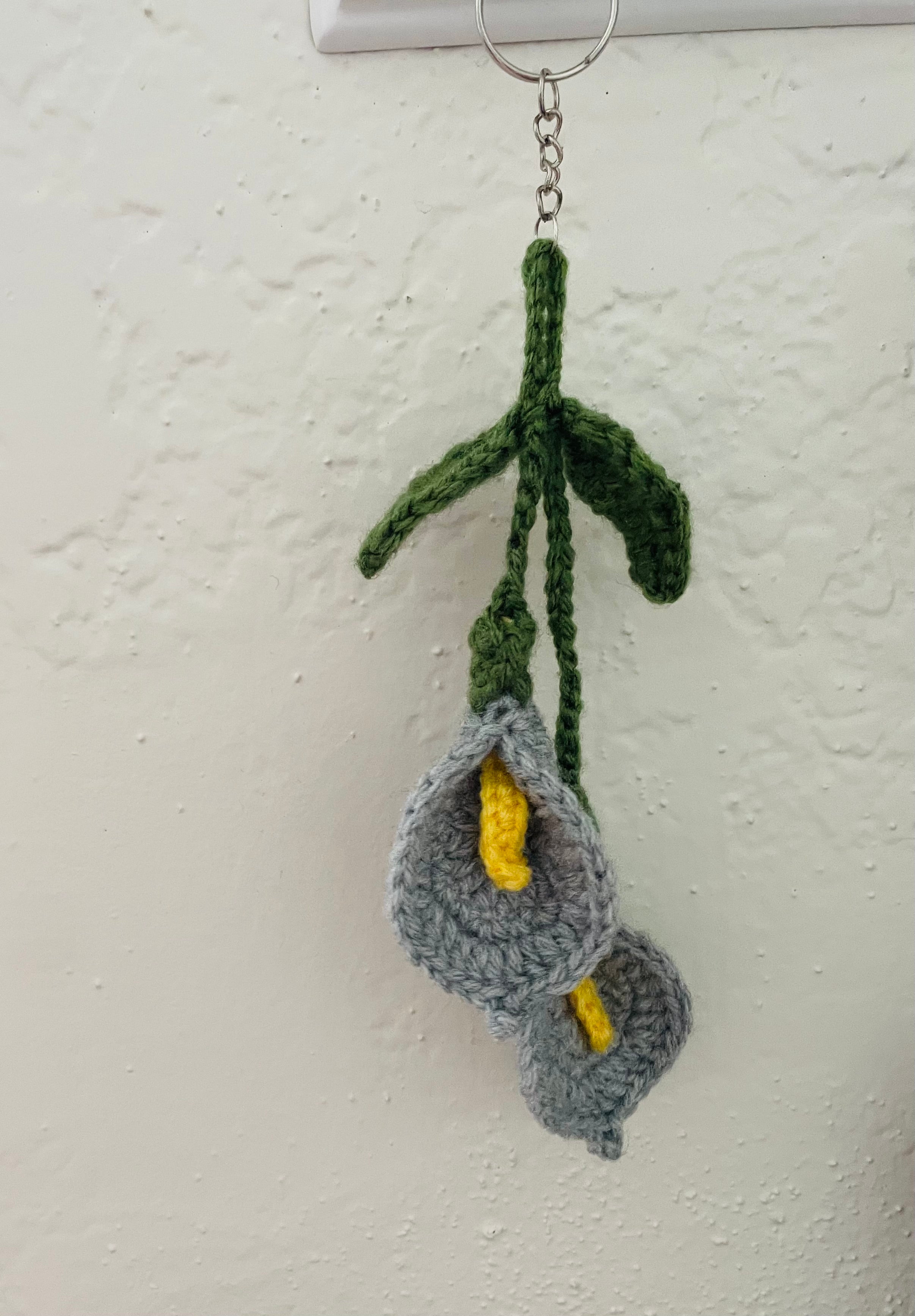 A handmade crocheted keychain featuring two gray calla lilies with yellow centers and green stems, hanging from a metal ring against a textured off-white wall.