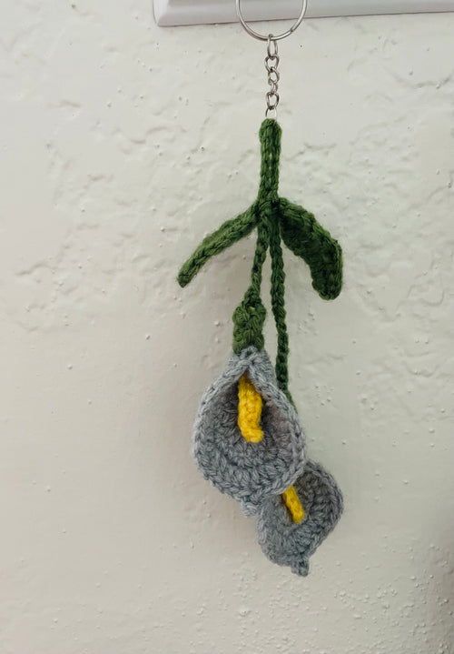 A handmade crocheted keychain featuring two gray calla lilies with yellow centers and green stems, hanging from a metal ring against a textured off-white wall.
