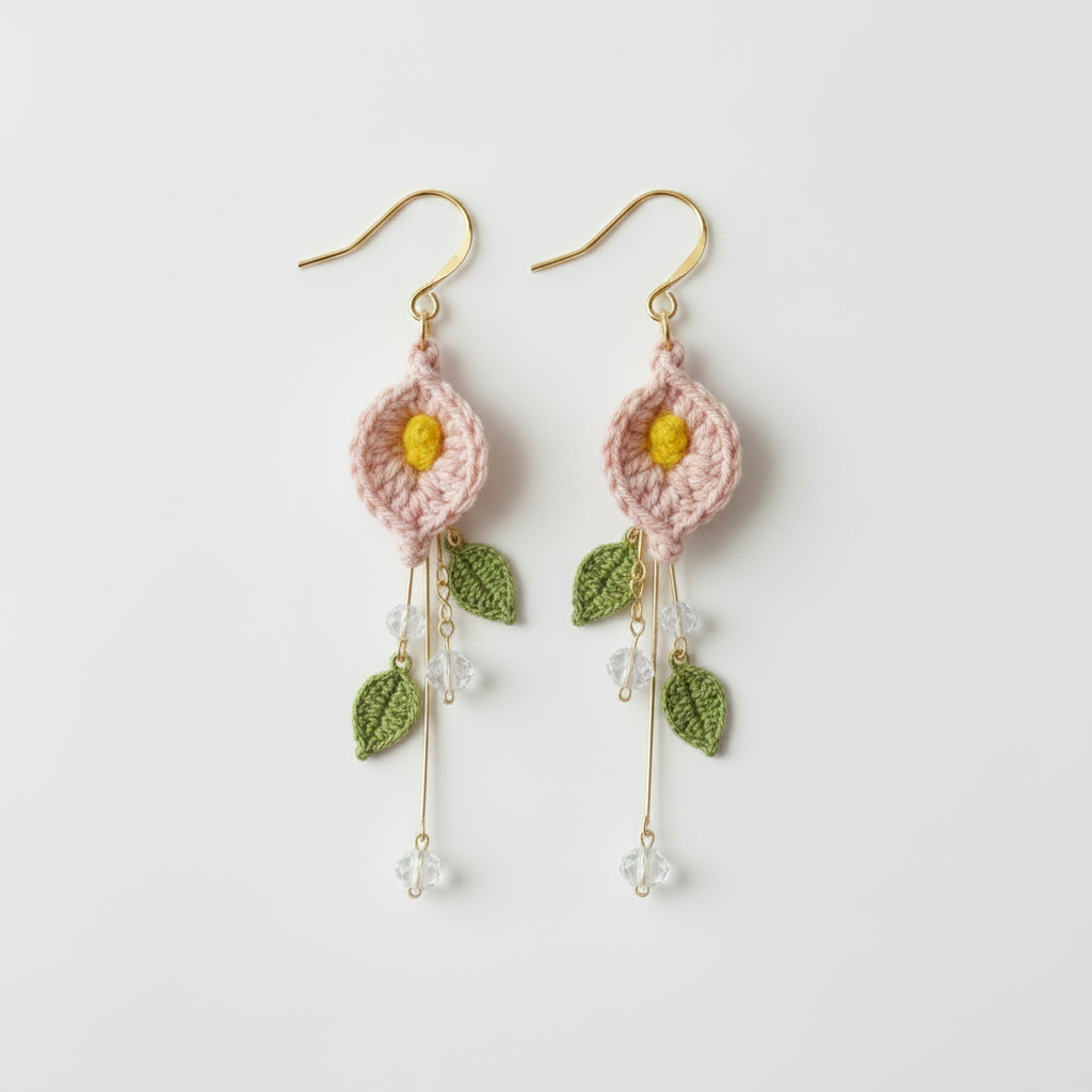 A pair of handmade crochet dangle earrings featuring light pink flower shapes with yellow centers, green leaves, and gold-tone hooks on a plain light gray background.
