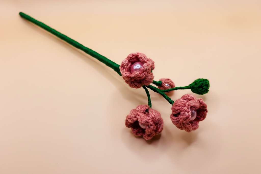 A handmade crocheted floral hair accessory featuring three pink flowers with pearl-like centers and green stems, displayed on a solid beige background.