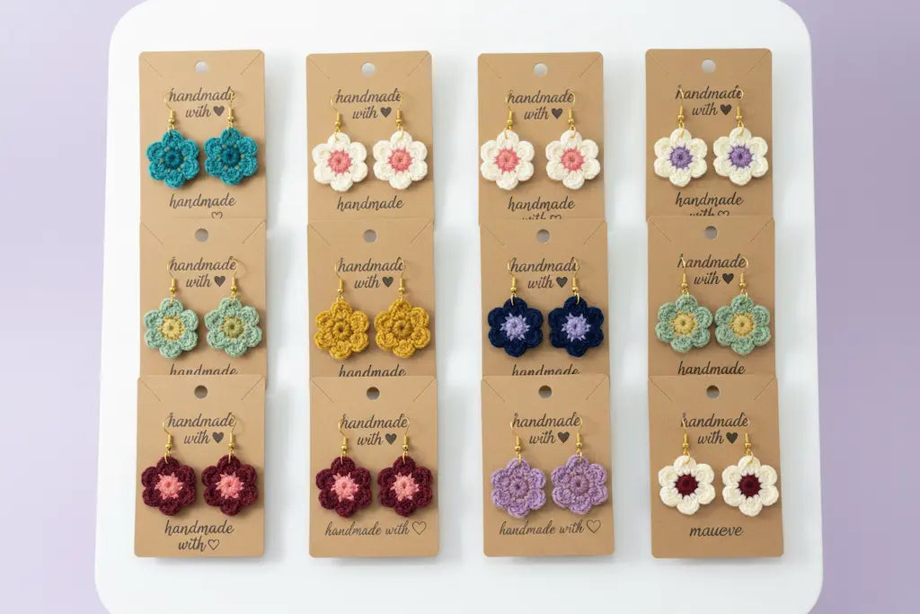 Six pairs of handmade floral stud earrings displayed on tan cardboard cards, arranged in two columns on a white stand against a soft lavender background.