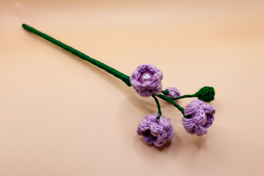 A handmade crochet flower stem featuring purple crocheted blossoms with a small pearl center, green stems, and a green leaf, set against a plain beige background.