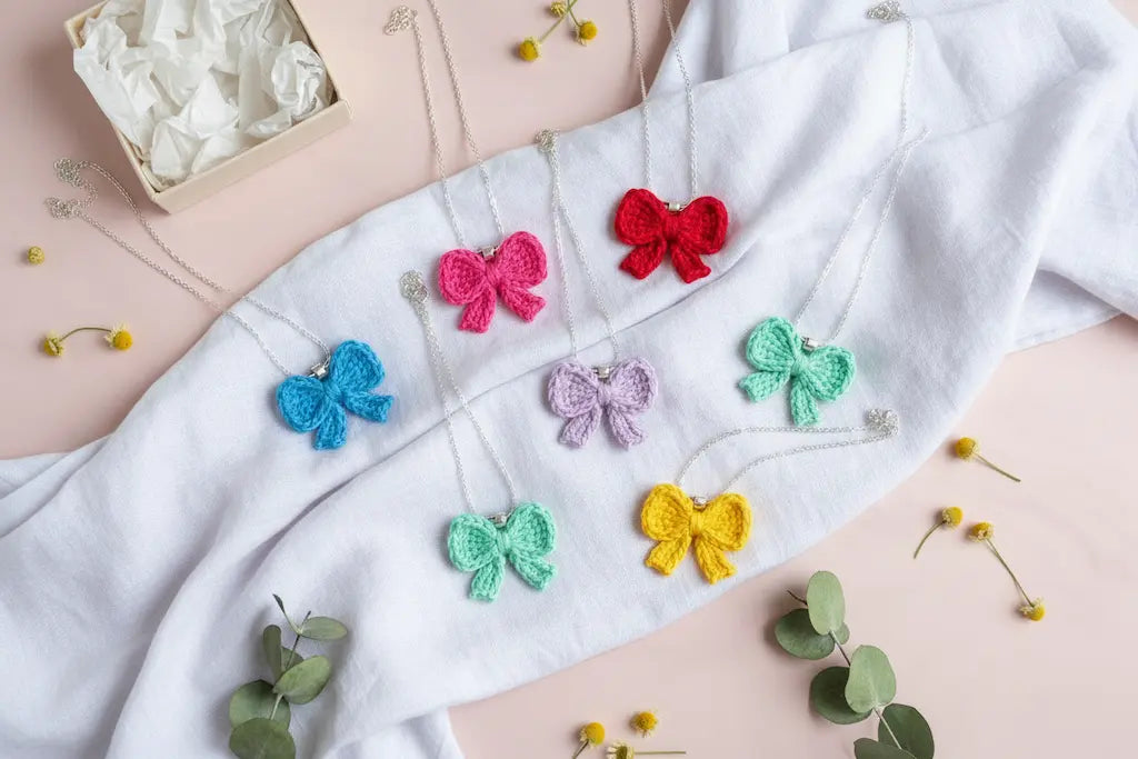 Six colorful crocheted bow necklaces in pink, red, blue, purple, green, and yellow, arranged on a white linen cloth over a light pink background with small eucalyptus sprigs and a gift box.