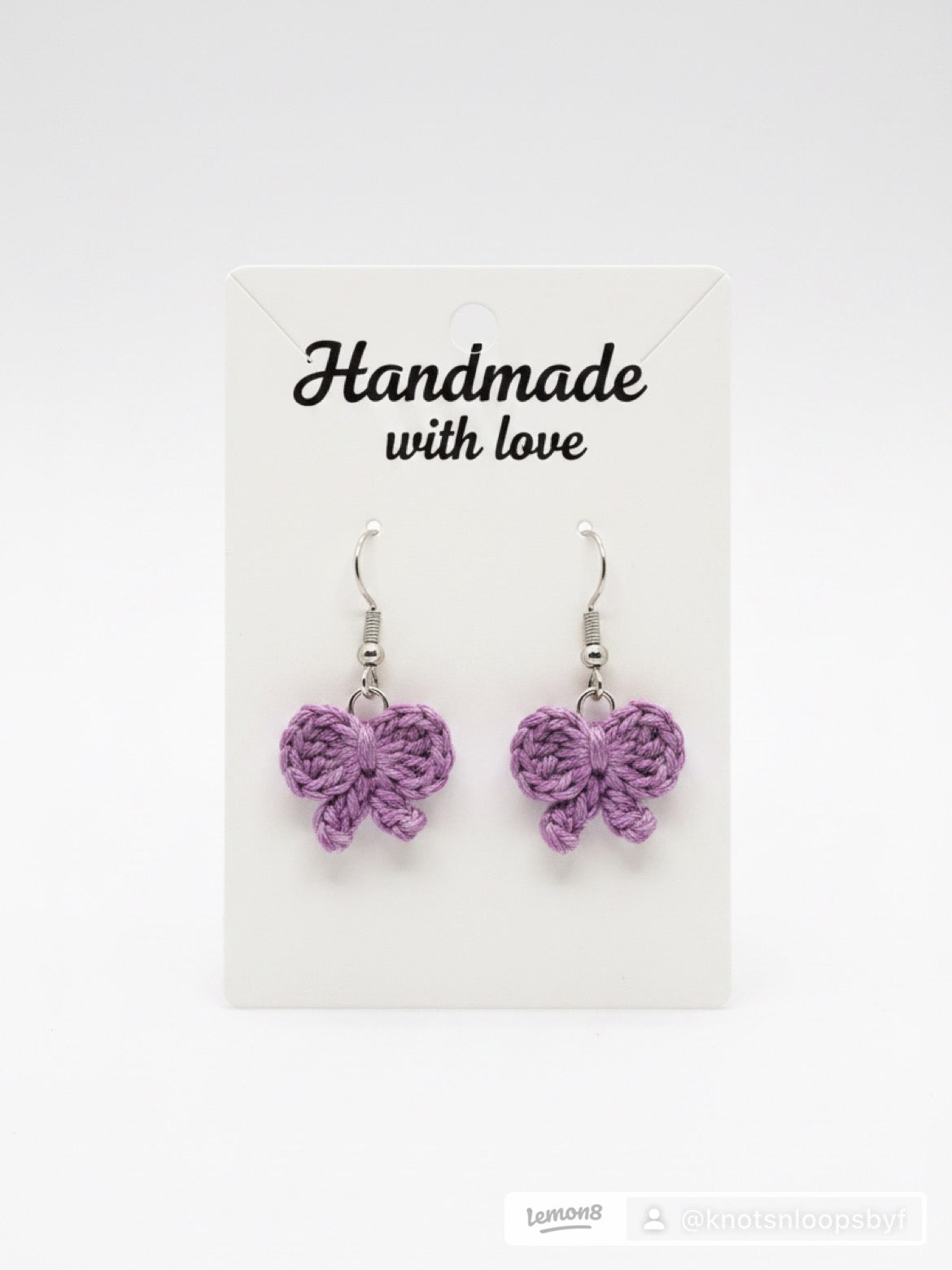 A pair of handmade purple crocheted bow dangle earrings on a white display card that reads 'Handmade with love'.