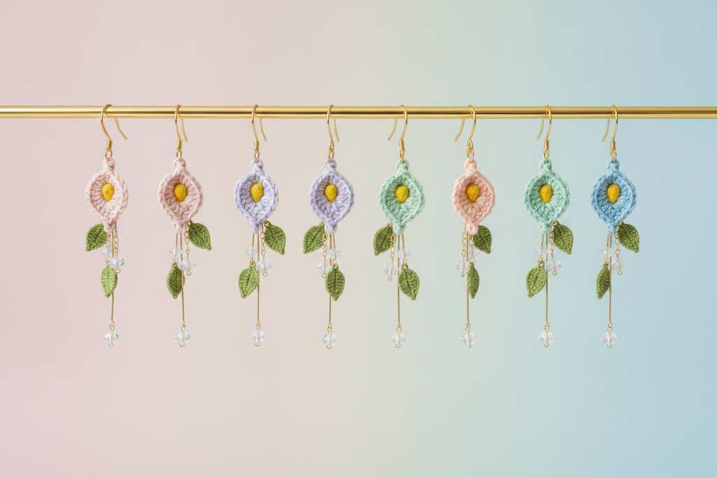 A set of nine handmade crocheted dangle earrings hanging from a gold rod against a soft pastel gradient background. The earrings feature floral motifs in pastel pink, lavender, mint green, and blue, with green leafy accents and dangling clear beads.
