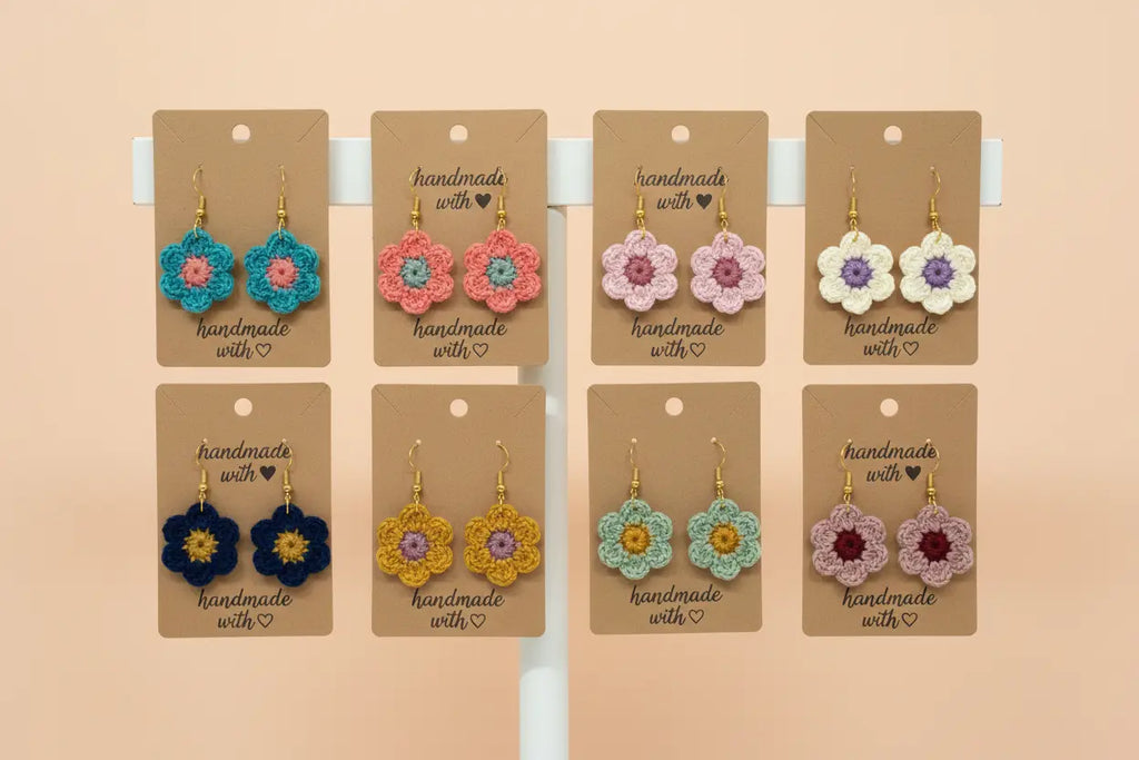 Eight pairs of handmade crocheted flower earrings displayed on brown kraft cards, arranged in two rows on a white stand against a warm beige background. The flowers feature various color combinations including turquoise, pink, lavender, yellow, and purple.