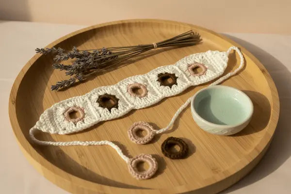 A crocheted white headband with four circular cutouts, displayed on a round wooden tray alongside dried lavender, small handmade ring accessories, and a small light blue dish.