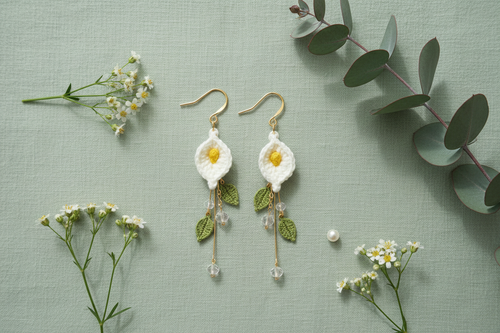 A pair of handmade crocheted daisy earrings with gold hooks, white petals, yellow centers, and green leaves, displayed on a textured sage green background with small white flowers and eucalyptus leaves.