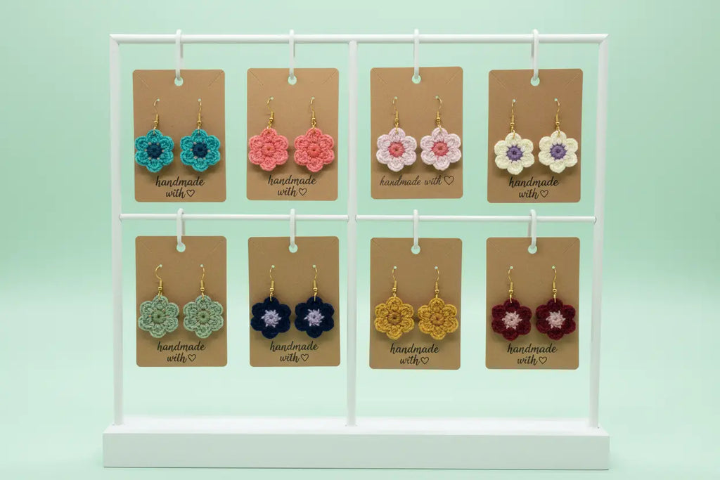 A white display stand holding eight handmade crocheted flower earrings on brown cards, set against a mint green background.