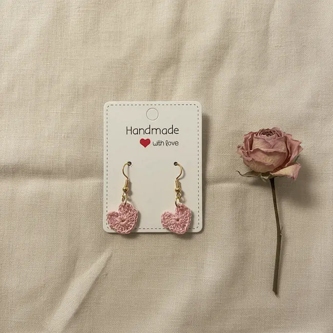 A pair of handmade pink crocheted heart dangle earrings on a white display card that reads 'Handmade with love', placed on a beige fabric background next to a dried pink rose.