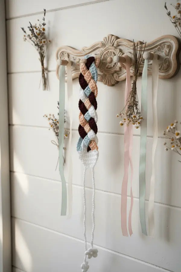 A crocheted braided hair accessory with a multi-colored braid in brown, beige, light blue, and white, hanging from a decorative wooden wall rack with pastel ribbons and dried flowers.