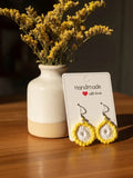 Yellow and white crocheted hoop earrings on a white 'Handmade with love' display card, set on a wooden table next to a ceramic vase with dried yellow flowers.