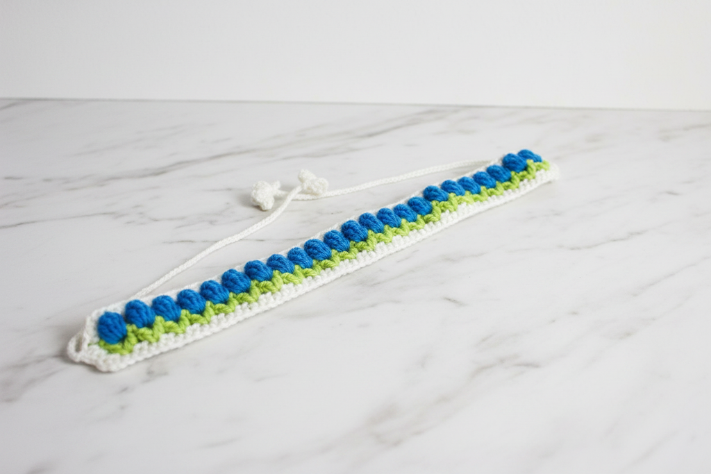 A handmade crochet hair claw clip made of white, blue, and lime green yarn, resting on a white marble surface.