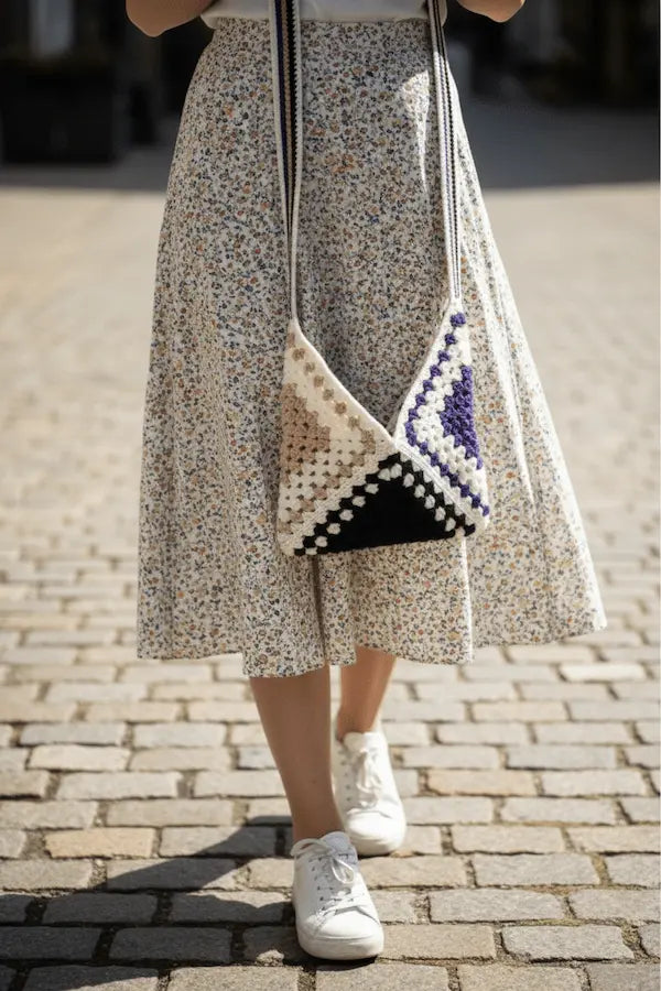 A person outdoors on a cobblestone street wearing a cream floral midi skirt and carrying a woven triangular shoulder bag with black, white, and purple sections.