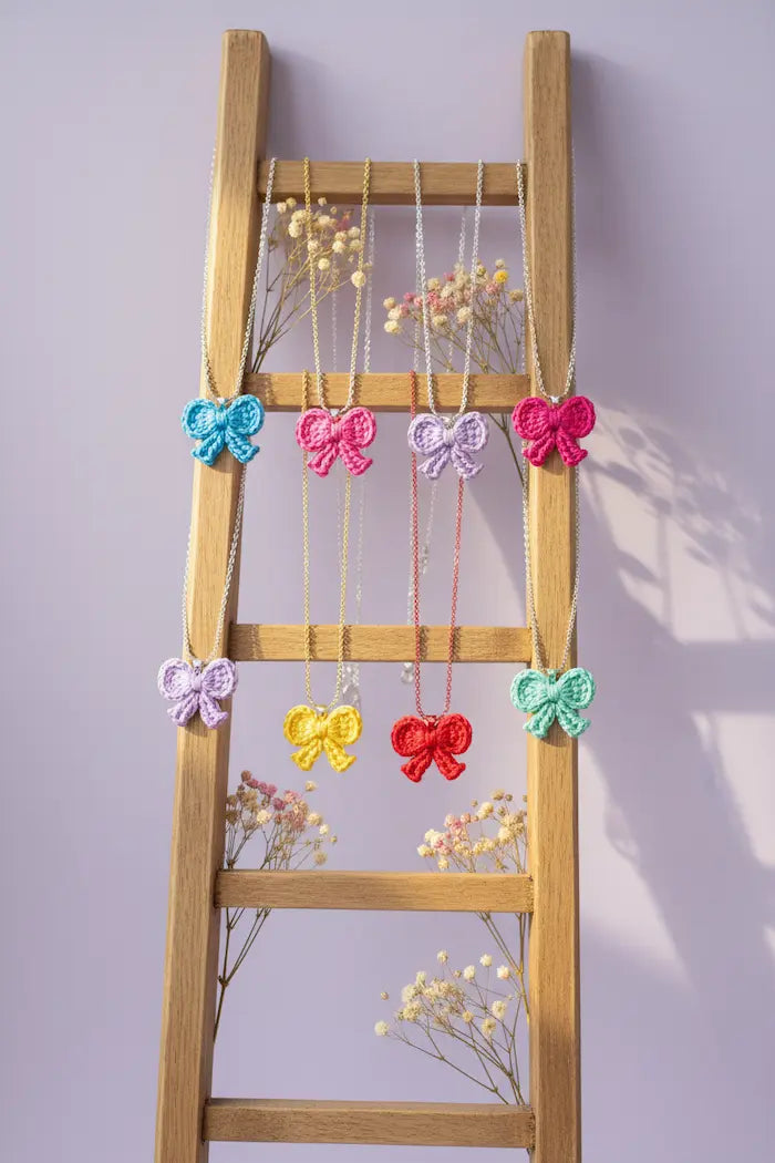 Six colorful crocheted bow necklaces in shades of blue, pink, purple, yellow, red, and mint green, hanging from a wooden ladder against a light lavender wall with dried baby's breath flowers.