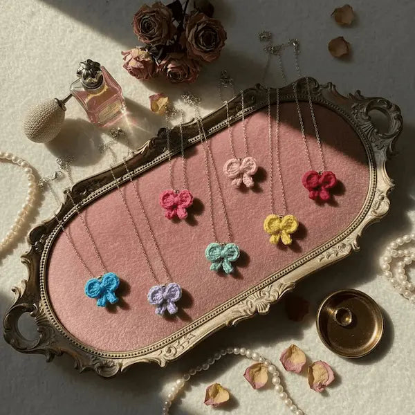 A styled flat-lay of nine knotted bow necklaces in various colors (blue, purple, green, pink, red, and light pink) hanging from a decorative gold-toned metal tray with a pink velvet lining. The tray is surrounded by vintage props including a perfume bottle, dried roses, pearls, and petals on a textured cream background.