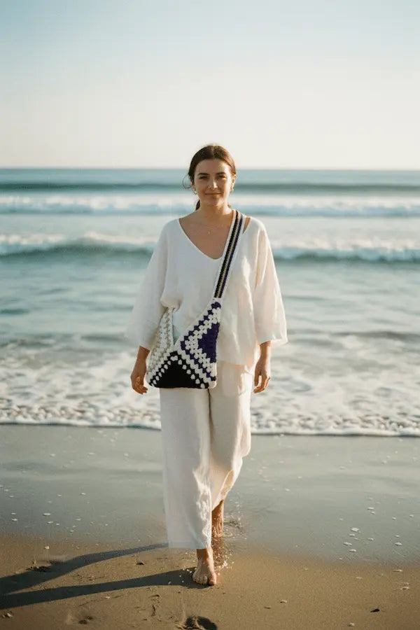 A woman walking on a sandy beach wearing a white two-piece lounge set and carrying a black and white patterned shoulder bag.