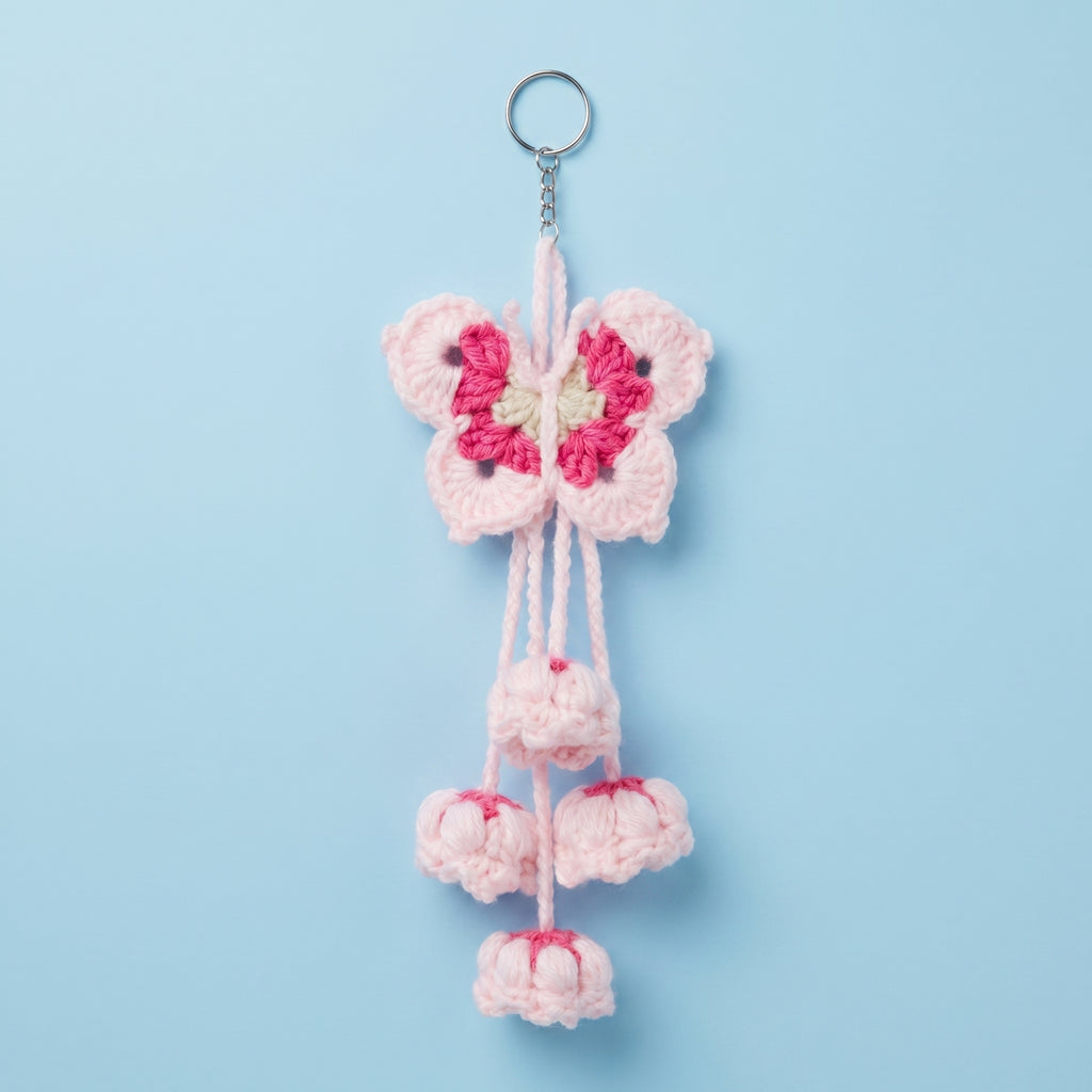 A crocheted cherry blossom keychain featuring a light pink butterfly with a bright pink and cream center, hanging from a silver metal key ring against a solid light blue background.