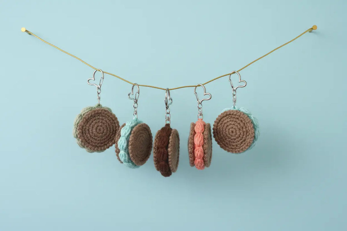 Five crocheted macaron-shaped earrings hang from a thin gold string against a light blue background. The earrings feature various macaron designs in brown, light blue, dark brown, peach, and mint green, each with silver metal hooks.