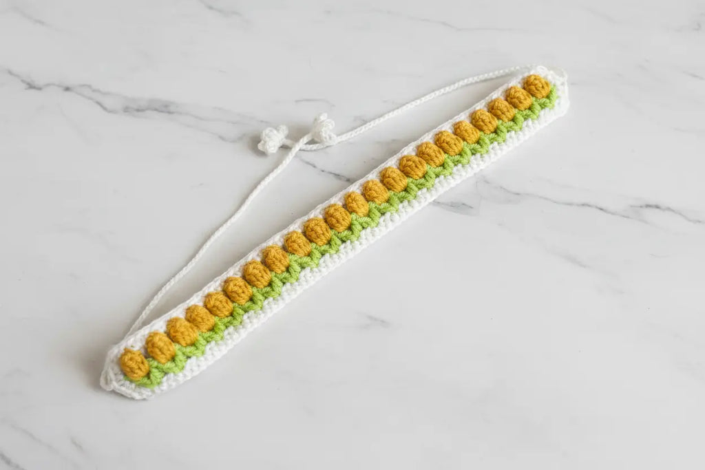 A handmade crocheted headband with a white base, a row of yellow flower-like stitches, and a row of green flower-like stitches, laid flat on a white marble surface.