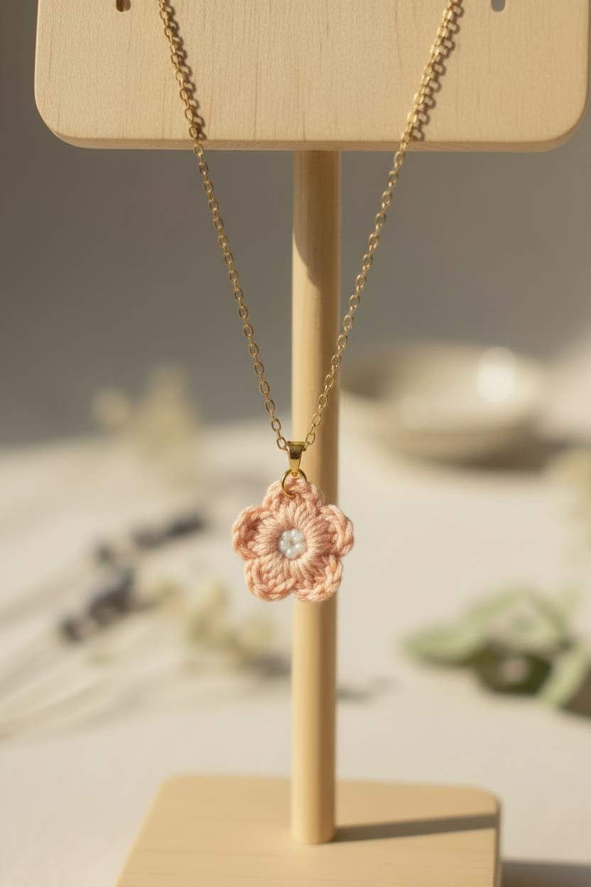 A gold chain necklace with a crocheted light pink flower pendant featuring a small white center, displayed on a light wood stand.