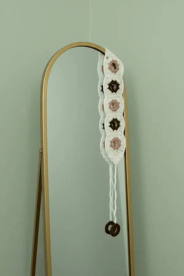 A white crocheted macrame wall hanging with pink and dark brown button accents, suspended from a gold-framed mirror against a pale green wall.