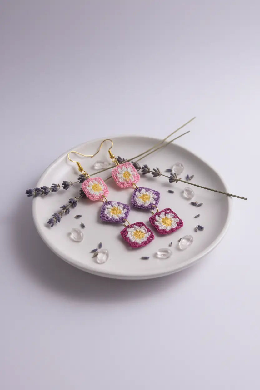A pair of handmade crochet flower earrings in pink and purple with yellow centers and gold hooks, displayed on a white ceramic plate with dried lavender sprigs and clear glass beads on a light gray background.
