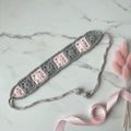 A crocheted hair claw clip in a gray and light pink granny square pattern, styled on a white marble background with a pink ribbon and dried bunny tail stems.