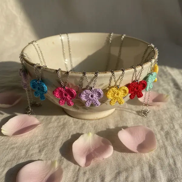 A ceramic bowl filled with silver chain necklaces, each featuring a crocheted butterfly charm in a different bright color including blue, pink, purple, yellow, red, and mint green. Pink flower petals are scattered on the linen surface around the bowl.