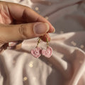 A close-up of a hand holding a pair of small pink crocheted heart dangle earrings with gold hooks against a soft pink satin fabric background.