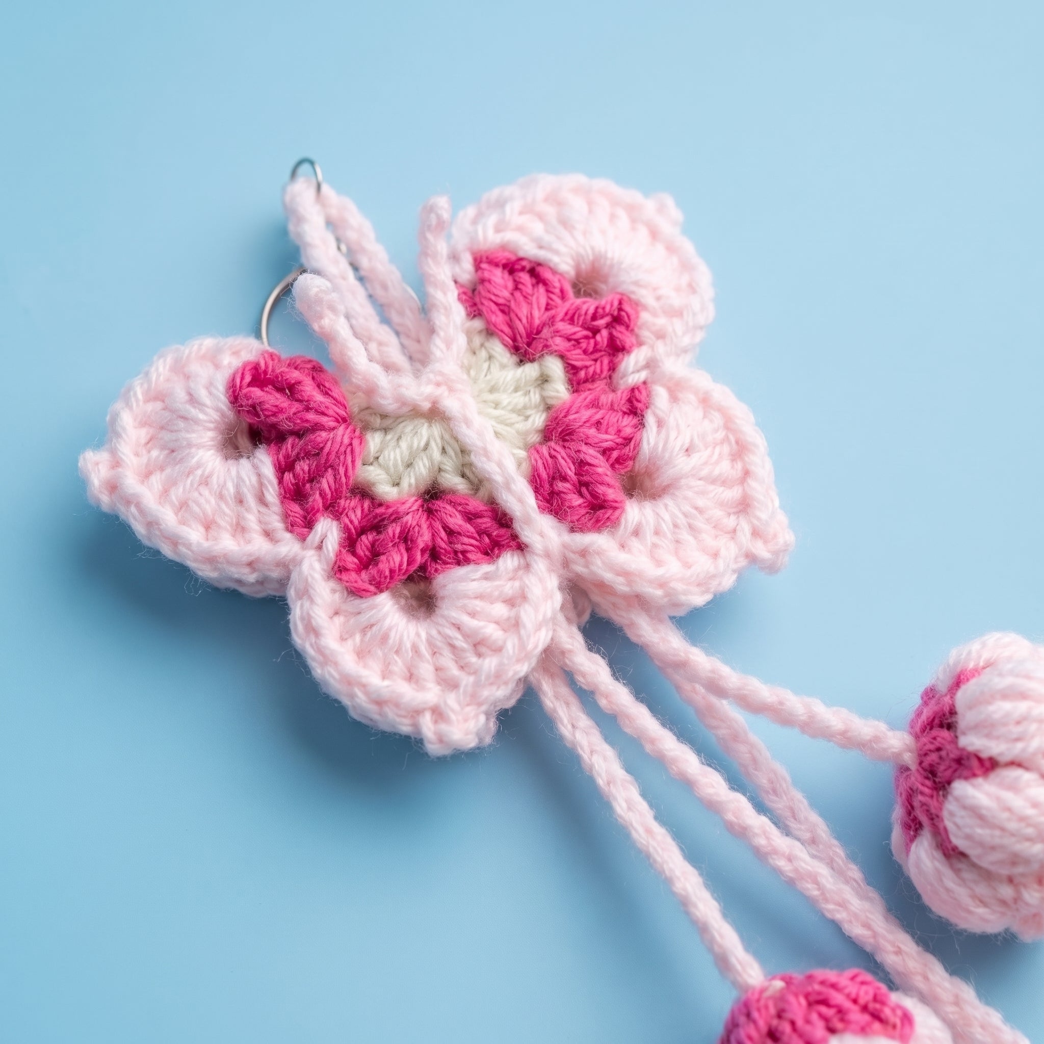 A crocheted flower-shaped hair clip in light pink, magenta pink, and cream, shown on a light blue background with its elastic bands visible.