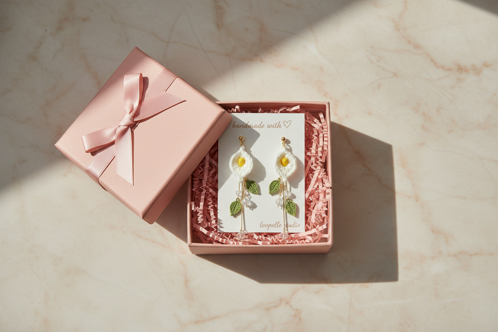A pink gift box with a satin ribbon bow sits beside an open pink presentation box containing a pair of dangling earrings shaped like flowers with green leaves, set on a light marble surface.