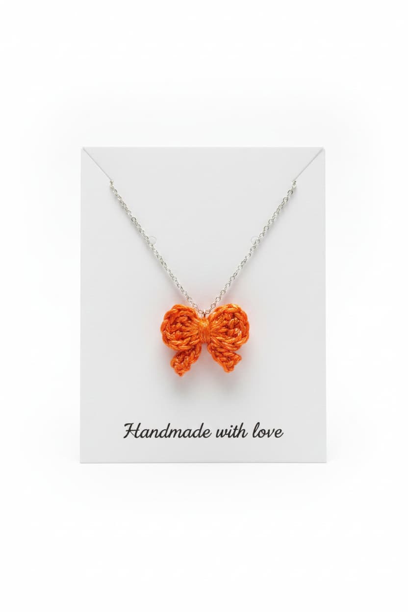 An orange crocheted bow pendant on a silver chain, displayed on a white backing card with the text 'Handmade with love' printed at the bottom.