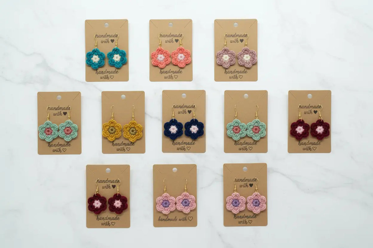 Nine pairs of handmade crocheted dangle earrings displayed on tan cards over a white marble background. The earrings feature colorful floral patterns in teal, coral, mauve, yellow, navy, mint, burgundy, and pink.