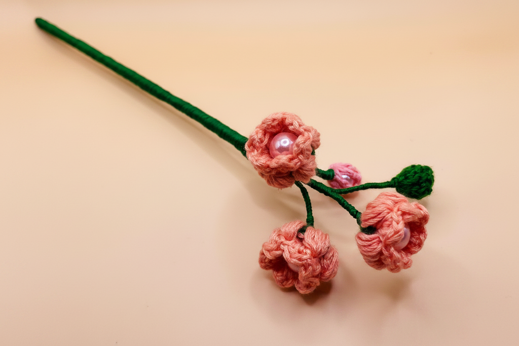 A handmade pink crocheted floral hair accessory with a green stem, three pink flower buds, and small pearl-like center accents, displayed on a light beige background.