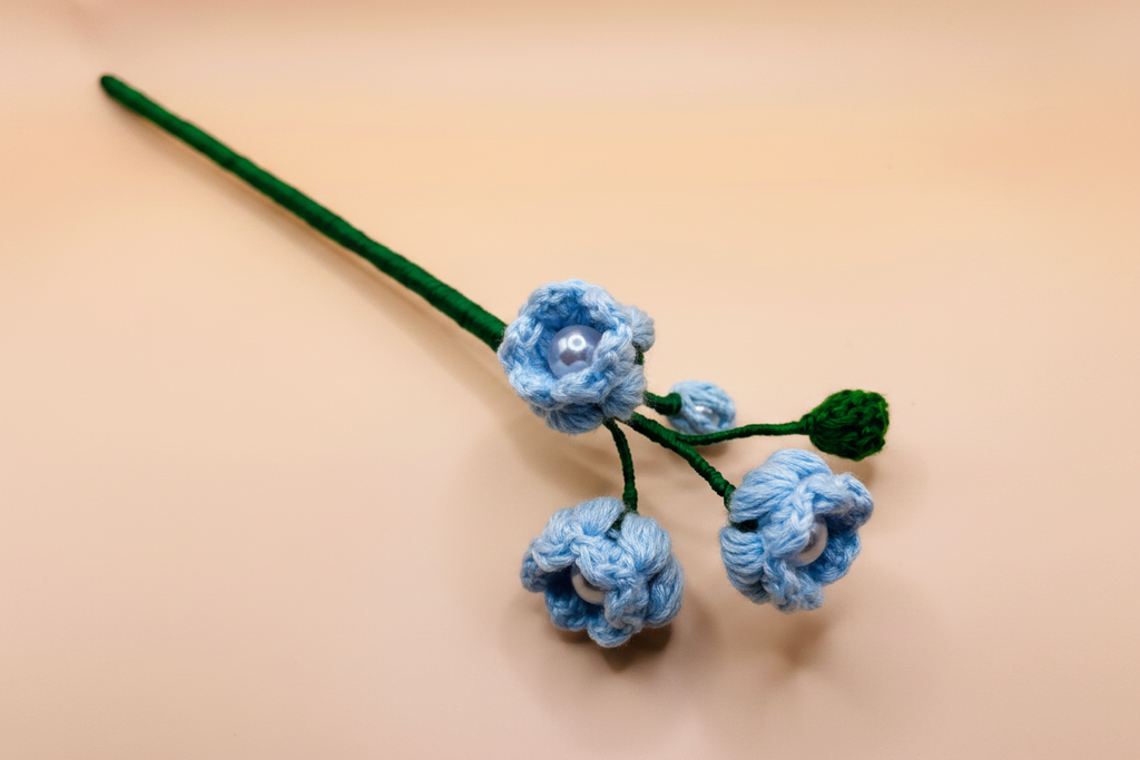 A handmade crocheted flower stem with three light blue flowers, green stems and leaves, and small pearl-like centers, displayed on a plain beige background.