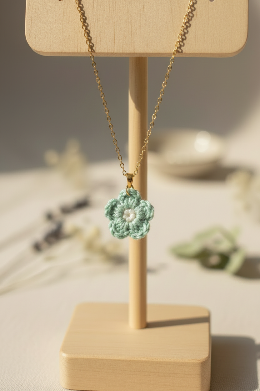 A gold chain necklace with a crocheted mint green flower pendant displayed on a light wood stand against a warm, softly blurred background.