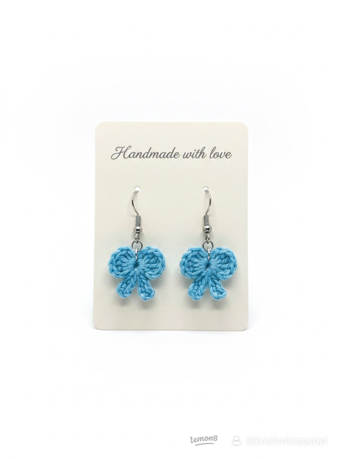 A pair of handmade light blue crocheted bow dangle earrings on a cream display card that reads 'Handmade with love'.