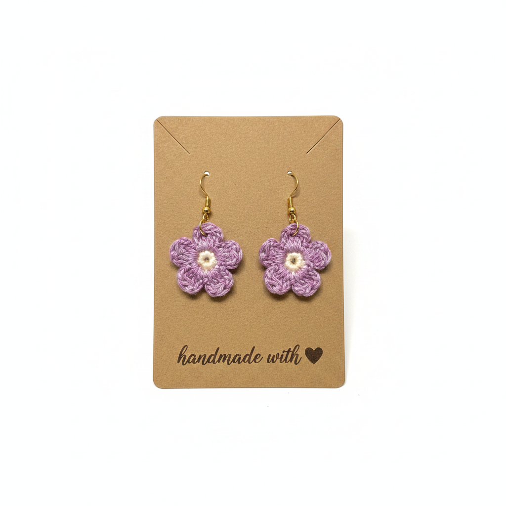 A pair of handmade lavender crocheted flower dangle earrings with gold hooks and white centers, displayed on a brown kraft paper card that reads 'handmade with heart'.