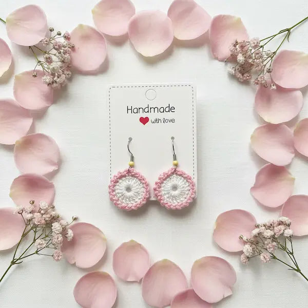 A pair of handmade crocheted dangle earrings in pink and white, displayed on a white card that reads 'Handmade with love'. The card is styled in a flat-lay arrangement with scattered pink petals and small white flowers on a white background.