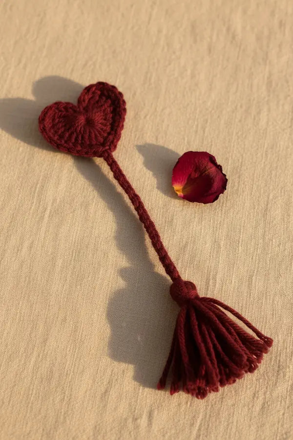 A dark red crocheted heart-shaped tassel accessory with a long cord and tassel, displayed on a textured beige surface next to a single red rose petal.