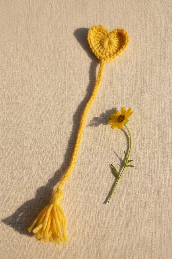 A yellow crocheted bookmark with a heart-shaped top and tassel end, styled on a textured beige background next to a small yellow flower.