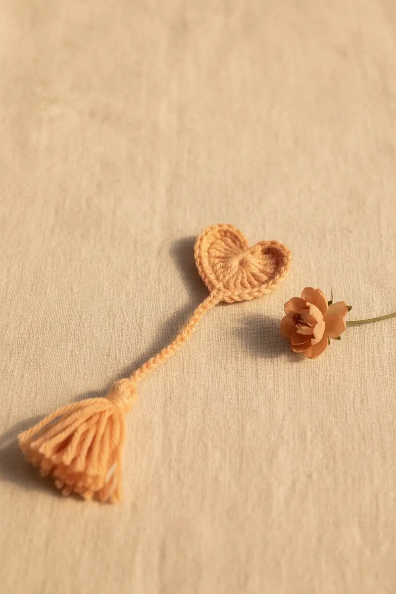 A small crocheted heart-shaped tassel accessory in a warm peachy-orange color, displayed on a textured beige fabric next to a dried flower.